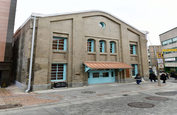 베이지색 벽돌 2층 건물 수원 구 부국원의 정면 전경, 파란 문과 구리색 캐노피